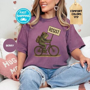 May include: Berry-colored t-shirt featuring a frog riding a bicycle and holding a sign that reads "RESIST." The frog wears a hat and backpack. The shirt has short sleeves and a relaxed fit. The image also includes a "Comfort Colors" tag.