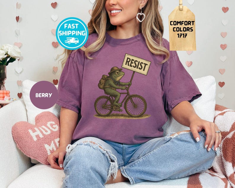 May include: Berry-colored t-shirt featuring a frog riding a bicycle and holding a sign that reads "RESIST." The frog wears a hat and backpack. The shirt has short sleeves and a relaxed fit. The image also includes a "Comfort Colors" tag.