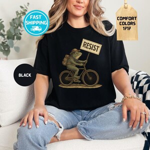 May include: Black t-shirt featuring a whimsical illustration of a frog riding a bicycle and holding a sign that reads "RESIST." The frog wears a hat and backpack. The shirt is labeled "Comfort Colors 1717."