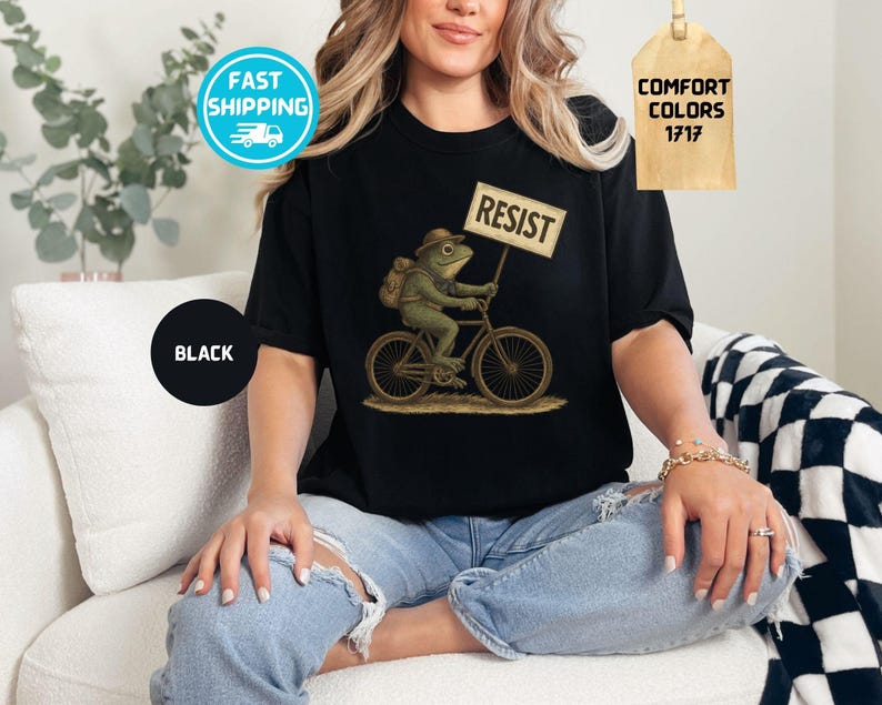 May include: Black t-shirt featuring a whimsical illustration of a frog riding a bicycle and holding a sign that reads "RESIST." The frog wears a hat and backpack. The shirt is labeled "Comfort Colors 1717."