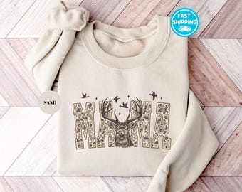 May include: Sand-colored sweatshirt with the word "MAMA" in a camouflage pattern, featuring a deer head graphic. The design includes flying birds and stars. The sweatshirt has a crew neck and long sleeves.