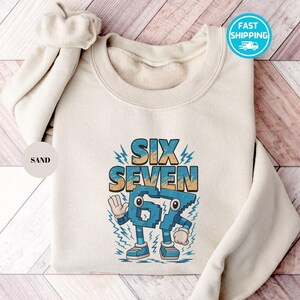 May include: A sand-colored sweatshirt with the words "SIX SEVEN" in a retro font. The number "67" is designed as a cartoon character with arms, legs, and shoes. The design is in shades of blue, teal, and orange. A "Fast Shipping" sticker is visible.