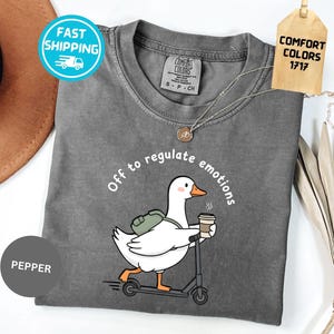 May include: A grey t-shirt featuring a white goose on a scooter, carrying a coffee cup and a backpack, with the text "Off to regulate emotions". The shirt has a Comfort Colors tag. The shirt is a Pepper color.