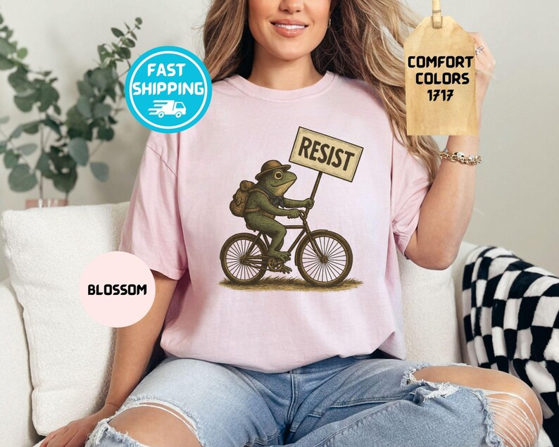 May include: Light pink t-shirt featuring a whimsical illustration of a frog riding a bicycle and holding a sign that reads "RESIST." The shirt also has the word "BLOSSOM" printed on it.