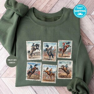 May include: Military green sweatshirt featuring six vintage-style postage stamp designs of rodeo riders on bucking horses. The sweatshirt has a crew neck and long sleeves. The text "MILITARY GREEN" is visible on the left side.