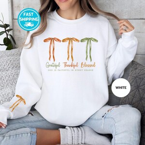 Pu&ograve; includere: Felpa bianca con fiocchi ad acquerello arancioni e verdi. Il testo recita "Grateful Thankful Blessed" con "GOD IS FAITHFUL IN EVERY SEASON" sotto. Un cerchio bianco con la parola "WHITE" si trova in basso a destra.