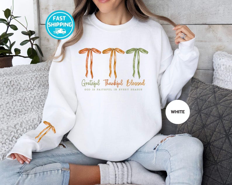 Pu&ograve; includere: Felpa bianca con fiocchi ad acquerello arancioni e verdi. Il testo recita "Grateful Thankful Blessed" con "GOD IS FAITHFUL IN EVERY SEASON" sotto. Un cerchio bianco con la parola "WHITE" si trova in basso a destra.