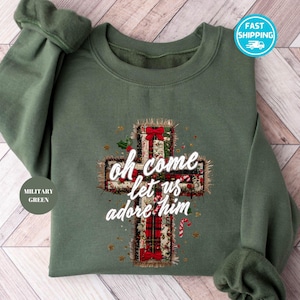 Peut inclure: Sweat-shirt vert militaire avec un motif de croix. La croix présente un motif à carreaux, des nœuds rouges, du houx et le texte "Oh come let us adore him". Le sweat-shirt a des manches longues et un col rond.