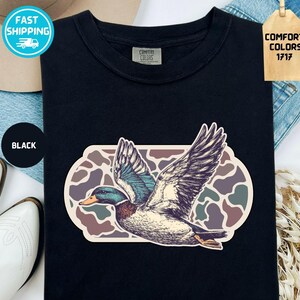 May include: Black t-shirt featuring a detailed illustration of a flying duck with outstretched wings. The duck has blue, green, and brown feathers, set against a camouflage-style background. The shirt has a Comfort Colors tag.
