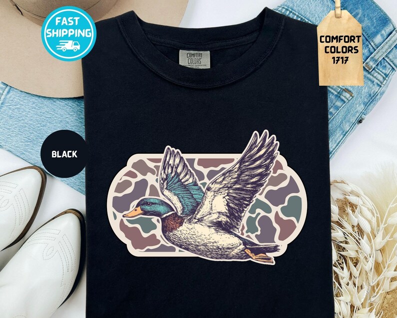 May include: Black t-shirt featuring a detailed illustration of a flying duck with outstretched wings. The duck has blue, green, and brown feathers, set against a camouflage-style background. The shirt has a Comfort Colors tag.