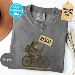 May include: A gray t-shirt with a graphic of a frog riding a bicycle and holding a sign that says "RESIST". The frog is wearing a hat and backpack. The shirt has a crew neck and short sleeves.