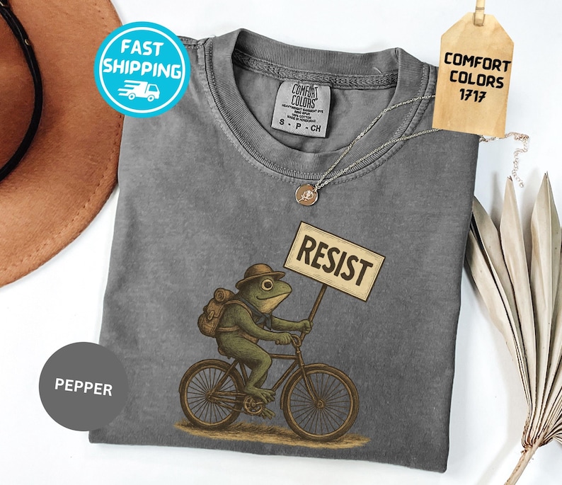 May include: A gray t-shirt with a graphic of a frog riding a bicycle and holding a sign that says "RESIST". The frog is wearing a hat and backpack. The shirt has a crew neck and short sleeves.