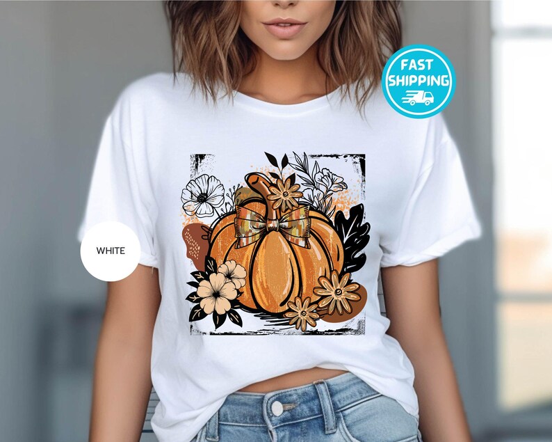 May include: White t-shirt featuring a vibrant orange pumpkin graphic with a plaid bow, surrounded by floral and leaf accents. The design is framed within a black border. A white circle with the word "WHITE" is on the left side of the shirt.