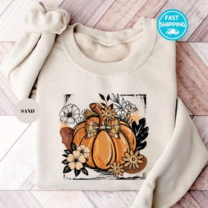 May include: Sand-colored sweatshirt featuring a vibrant orange pumpkin with a plaid bow, surrounded by floral and leaf illustrations. The design is framed within a black rectangle. The text "FAST SHIPPING" is on the upper right.