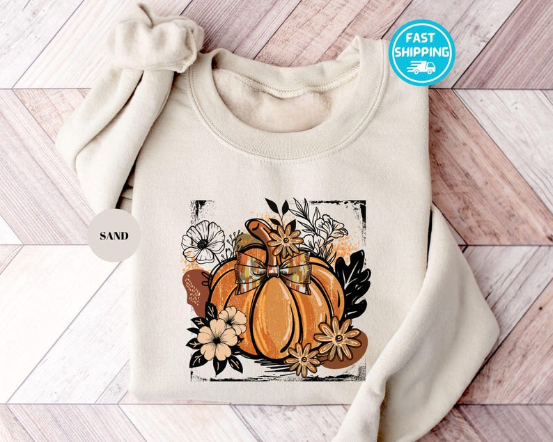 May include: Sand-colored sweatshirt featuring a vibrant orange pumpkin with a plaid bow, surrounded by floral and leaf illustrations. The design is framed within a black rectangle. The text "FAST SHIPPING" is on the upper right.