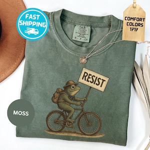 May include: Moss green t-shirt featuring a frog riding a bicycle and holding a sign that reads "RESIST." The frog wears a hat and backpack. The shirt has a Comfort Colors tag and a Fast Shipping sticker.