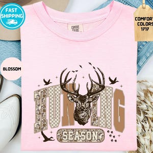 May include: A pale pink t-shirt featuring a deer head design and the text "HUNTING SEASON". The graphic includes a deer head, birds, and camouflage-style lettering. The shirt is a Comfort Colors brand.