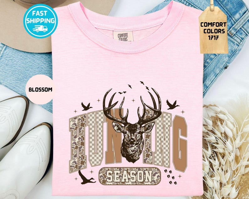 May include: A pale pink t-shirt featuring a deer head design and the text "HUNTING SEASON". The graphic includes a deer head, birds, and camouflage-style lettering. The shirt is a Comfort Colors brand.