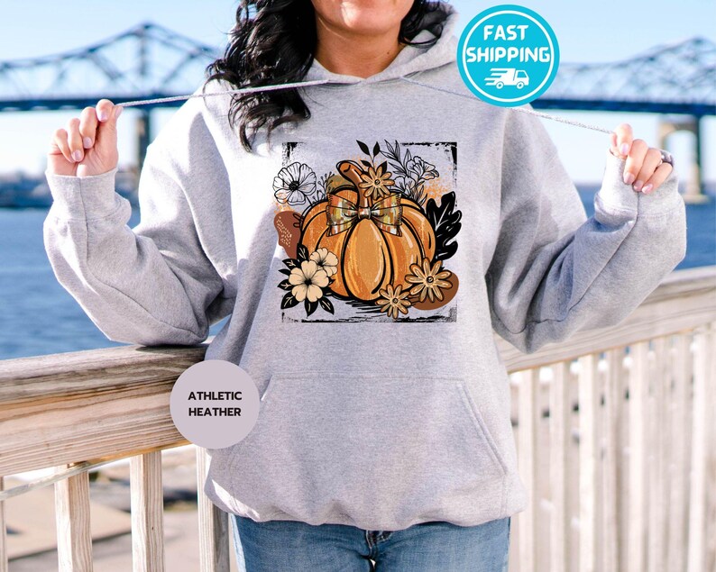 May include: Athletic heather gray hoodie with a pumpkin graphic. The pumpkin is orange with a bow and surrounded by flowers and leaves. The hoodie has a front pocket and a drawstring hood.
