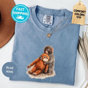 May include: Blue jean colored Comfort Colors t-shirt featuring a graphic of two monkeys hugging an orange stuffed animal. The shirt has a crew neck and short sleeves. A gold necklace is draped across the shirt.