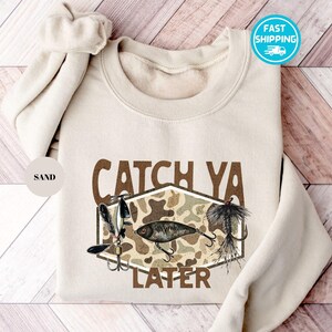 Puede incluir: Sudadera de color arena con el texto "CATCH YA LATER" y gráficos de señuelos de pesca. El diseño incluye un patrón de camuflaje y varios señuelos de pesca en tonos plateados, negros y marrones. La sudadera tiene cuello redondo y mangas largas.