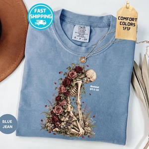 May include: A blue t-shirt featuring a graphic of a skeleton kneeling amidst red roses and greenery. The text Ezekiel 37:1-10 is printed in white next to the skeleton. The shirt has a Comfort Colors brand label on the inside collar.