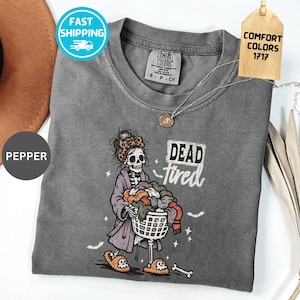 May include: A pepper-colored t-shirt featuring a graphic of a skeleton wearing a leopard print headband, a purple robe, and ghost-themed slippers while holding a laundry basket. The text "DEAD tired" is printed next to the skeleton.