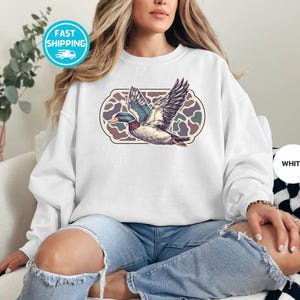 Peut inclure: Un sweat-shirt blanc avec un canard volant dans un motif g&eacute;om&eacute;trique. Le canard est bleu et vert avec une poitrine blanche. Le motif est brun, vert et blanc.