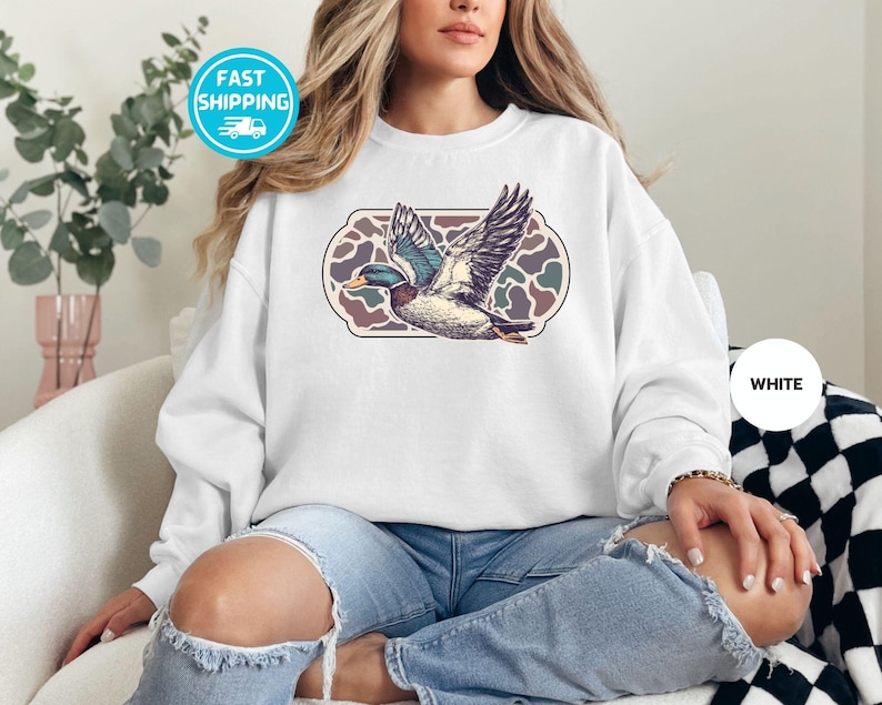 Peut inclure: Un sweat-shirt blanc avec un canard volant dans un motif g&eacute;om&eacute;trique. Le canard est bleu et vert avec une poitrine blanche. Le motif est brun, vert et blanc.