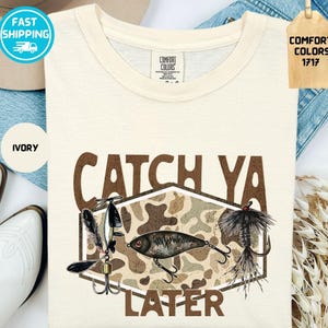 Puede incluir: Camiseta color marfil con la frase "CATCH YA LATER" en marrón. El diseño presenta señuelos de pesca y un patrón de camuflaje. La camiseta es de material Comfort Colors 1717.