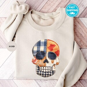 Könnte beinhalten: Ein sandfarbenes Sweatshirt mit einem Patchwork-Schädel-Design. Der Schädel weist verschiedene Stoffmuster auf, darunter blaues Karomuster, Orange und Weiß. Das Sweatshirt hat einen Rundhalsausschnitt und lange Ärmel. Der Text "SAND" befindet sich in einem Kreis.