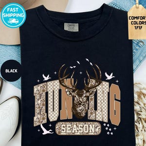 May include: Black t-shirt with a deer graphic and the words "HUNTING SEASON". The design features a deer head with antlers, camouflage and checkered patterns, and flying birds. The shirt is labelled "Comfort Colors 1717".