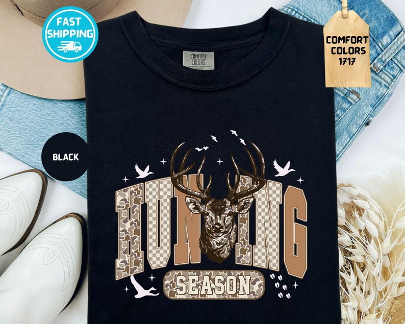 May include: Black t-shirt with a deer graphic and the words "HUNTING SEASON". The design features a deer head with antlers, camouflage and checkered patterns, and flying birds. The shirt is labelled "Comfort Colors 1717".