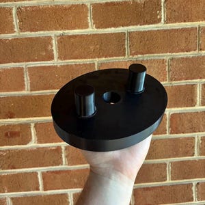 May include: A black, circular object with two cylindrical posts and a central hole. The object is held in a hand against a brick wall background. The object appears to be made of a solid, possibly plastic, material.