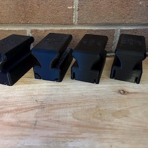 Puede incluir: Cuatro objetos negros y rectangulares con un diseño curvo están dispuestos sobre una superficie de madera. Los objetos tienen una ranura en un lado y una parte superior ligeramente inclinada. El fondo presenta una pared de ladrillos.