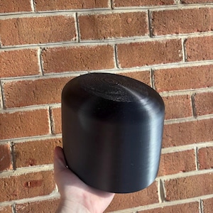 May include: A black, cylindrical object with a rounded top. The object has a smooth, matte finish and is held against a brick wall. The top of the object has a circular pattern.