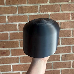 May include: A solid black cylindrical object with a rounded top. The object is held in front of a brick wall. The top of the cylinder has a textured, circular pattern.