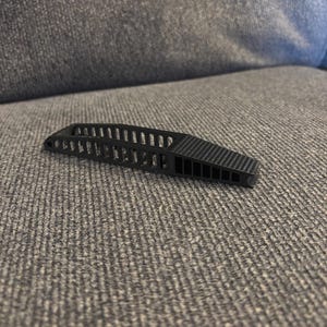 May include: A black, geometric object with a slotted design. The object has a curved shape with a series of parallel lines and a textured, angled section at one end. It appears to be made of a rigid material.