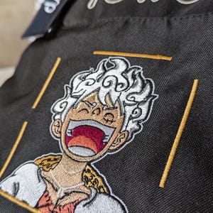 May include: Black backpack with a white and yellow embroidered image of a cartoon character with white hair and a red mouth. The backpack has the word "TOITO" embroidered on the front in white.