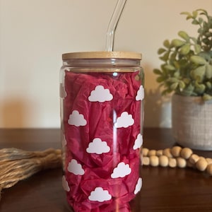 May include: A clear glass tumbler with white cloud decals, a bamboo lid, and a clear glass straw. The tumbler is filled with bright pink tissue paper. The tumbler is approximately 6 inches tall.