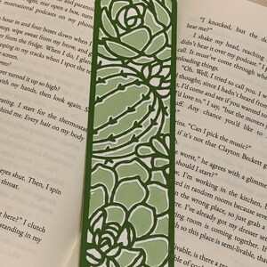 May include: A green bookmark with a cut-out design of a succulent plant. The bookmark is made of paper and has a white background. The bookmark is a decorative item for book lovers.