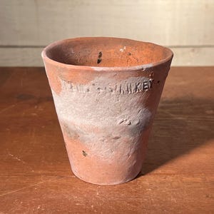 May include: A small, weathered terracotta flower pot with a slightly flared shape. The pot has a faded, aged appearance with visible text that reads "SANKEY". The pot is unglazed and has a rough texture, showing signs of use and age.