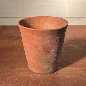 May include: A weathered, terracotta-colored clay pot with a slightly flared rim. The pot has a cylindrical shape with a textured surface and a band of embossed text near the top. The pot sits on a wooden surface.