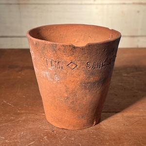 May include: A weathered, terracotta-colored flower pot with a slightly tapered shape. The pot has a rough texture and visible wear, with the text "T.M. SAHNE" etched into the side. The rim is uneven, adding to its rustic charm.