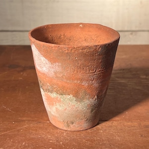 May include: A small, weathered terracotta pot with a cylindrical shape. The pot has a faded orange hue with white and green patches. The word "NUTSA" is embossed on the side. Ideal for small plants or as a decorative item.