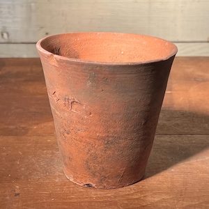 May include: A weathered, terracotta-colored flower pot with a slightly flared rim. The cylindrical pot has a textured surface and a small, embossed marking on the side. The pot is empty and sits on a wooden surface.