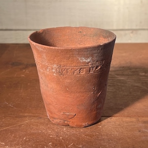 May include: A weathered, terracotta-colored clay pot with a slightly flared rim. The pot has a cylindrical shape with a textured surface and a band of embossed text near the top. The pot sits on a wooden surface.