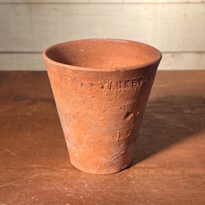 May include: A small, terracotta-colored clay pot with a slightly flared shape. The word "HIKEY" is stamped around the rim. The pot has a rustic, aged appearance, with visible texture and imperfections.
