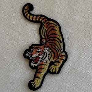 May include: An embroidered tiger patch in brown and gold with a black outline. The tiger is facing left and has its mouth open in a roar.