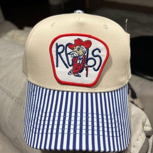 Puede incluir: Gorra de béisbol beige con una visera a rayas azules y blancas. La gorra presenta un parche con la palabra "REBS" y una imagen de dibujos animados de un hombre con un atuendo y sombrero rojos.